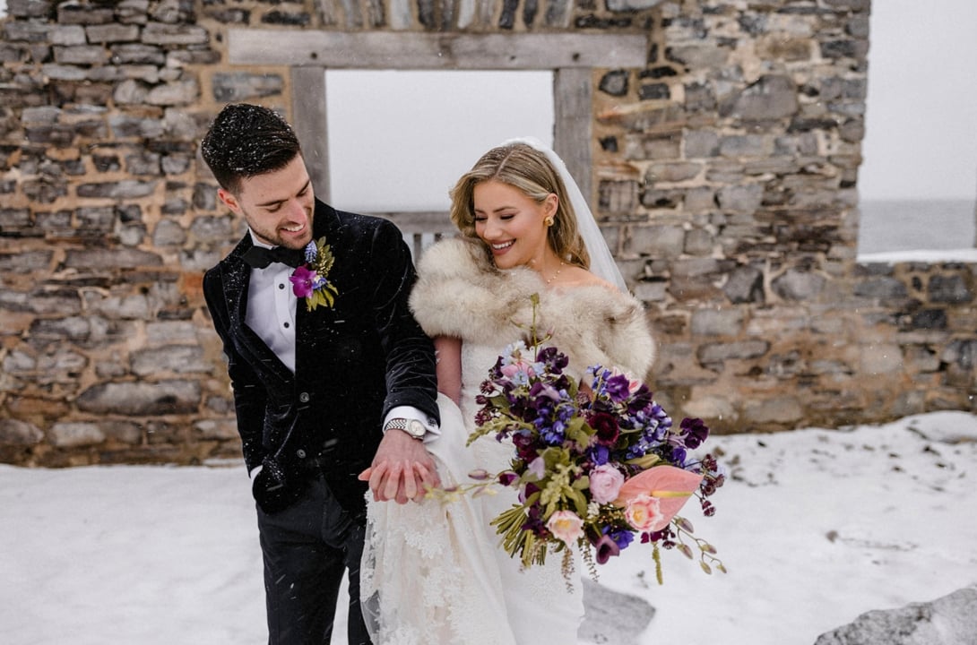 Smiling bride and groom holding hands in front of a stone house.